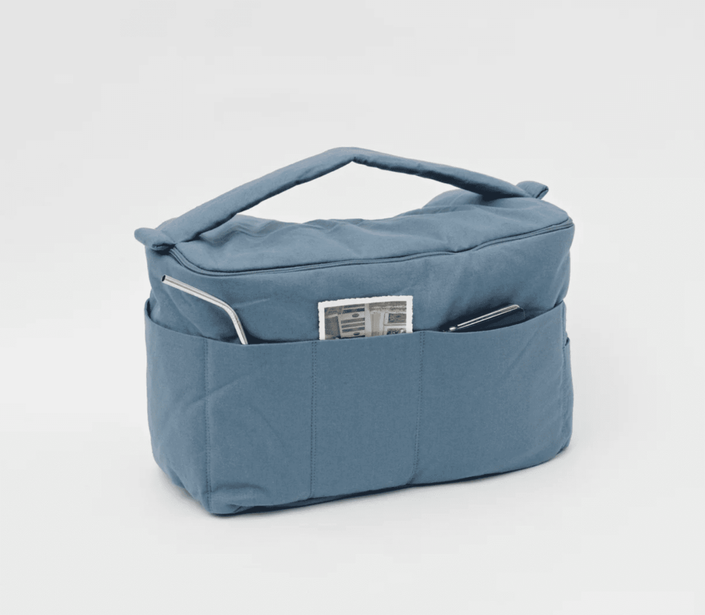 Eine hellblaue kompakte Stofftasche, die sich in Reichweite am Bettrahmen fixieren lässt. Mit Fächern außen, in denen Dinge stecken wie ein Strohhalm, Fotos und ein Handy.