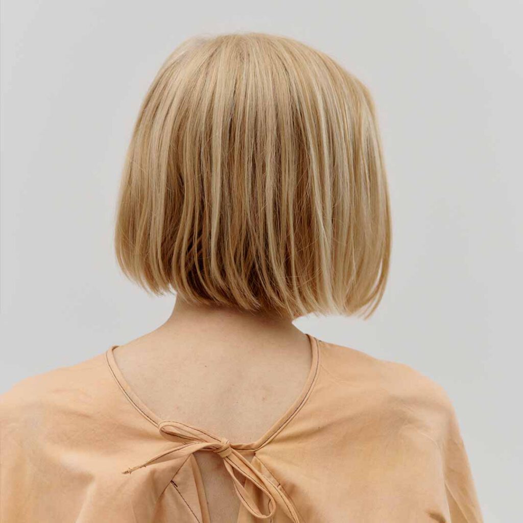 Hinterkopf und Schulterbereich einer blonden Frau mit Pagenschnitt. Das Hemd ist apricotfarben und mit einem Band im Nacken geschlossen.